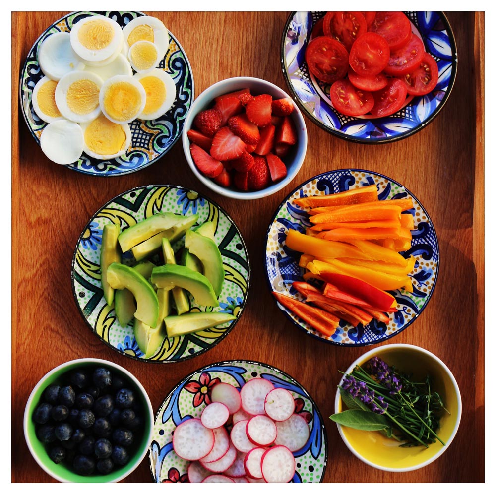 Acht Schüsseln, die mit Eiern, Erdbeeren, Tomaten, Avocados, Karotten, Heidelbeeren, Radieschen und Lavendel gefüllt sind.