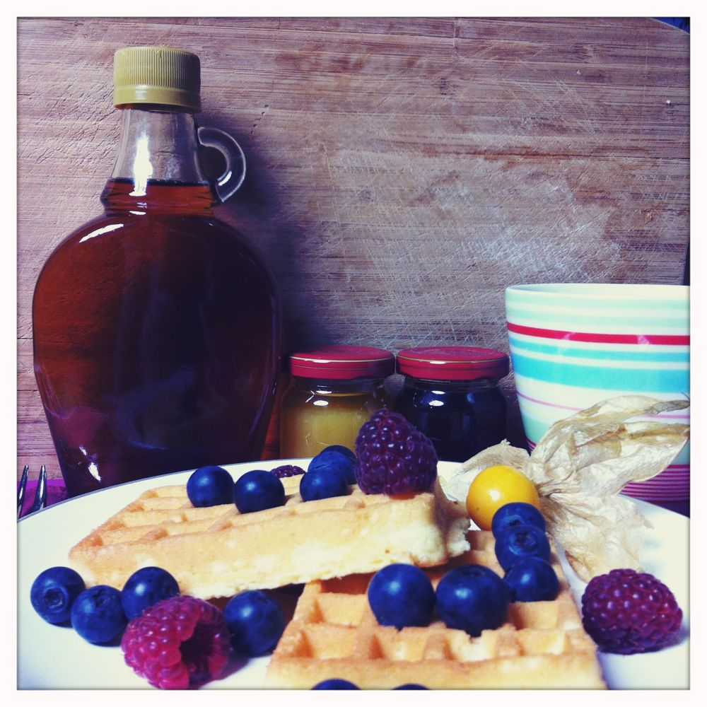 Ein Foto von Waffeln mit Waldbeeren, im Hintergrund ist eine Flasche Sirup, zwei Marmeladegläser und eine leere Tasse.